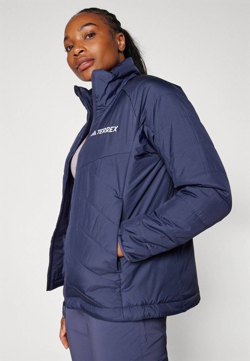 Veste en duvet marine avec un col montant, fermeture éclair et texture matelassée. Présente un logo sur la poitrine et des poches latérales. Design léger.