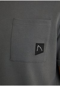 Camicia grigia a maniche corte realizzata in tessuto testurizzato, dotata di una tasca sul lato sinistro del petto e di un'etichetta quadrata nera con un design bianco.