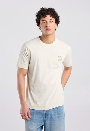 Jonge man met krullend haar, gekleed in een effen lichtbeige t-shirt met een klein bloemmotief op de borst en een blauwe spijkerbroek, staand tegen een effen achtergrond.