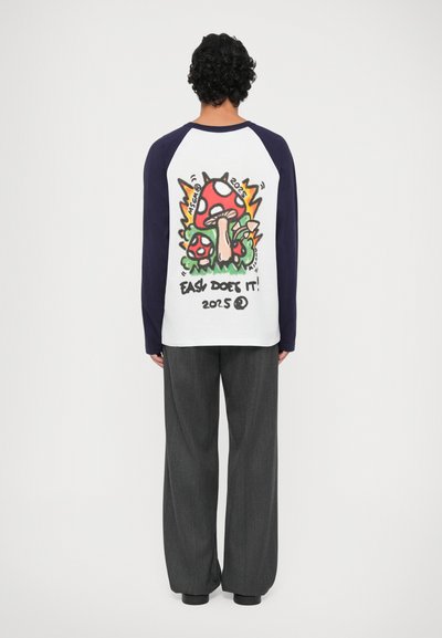 Chemise à manches longues avec un corps blanc et des manches bleu marine, arborant un graphisme coloré de champignons et de texte au dos. Associée à un pantalon large gris.
