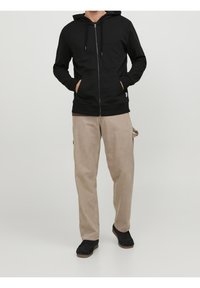 Černá mikina na zip s kapucí na šňůrku, klokankové kapsy a béžové kalhoty z měkké tkaniny, doplněné černými botami.