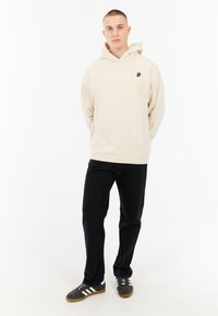 Crèmekleurige hoodie met een voorzak en een klein groen logo op de linkerborst; gecombineerd met zwarte broek en grijze sneakers.
