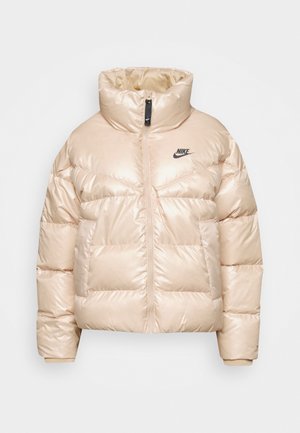 Puffa jakas gaiši bežā krāsā, izgatavota no spīdīga materiāla. Tai ir augsts apkakle, rāvējslēdzēja aizdare un melns Nike logo uz krūšu daļas.