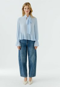 Frau steht und trägt eine hellblaue Bluse mit langen Ärmeln und vorderer Schlaufe, weite blaue Jeans und weiße spitze Schuhe vor weißem Hintergrund.