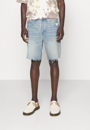 Hellblaue Jeansshorts mit ausgefranstem Saum, kombiniert mit einem Batik-T-Shirt und beigen Slip-On-Schuhen mit braunen Akzenten.