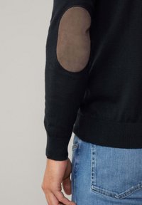 Czarny sweter z teksturowymi brązowymi łatkami na łokciach, prążkowanymi mankietami i dołem; w zestawieniu z jasnoniebieskimi jeansami. Skupienie na materiałach i szczegółach wzornictwa.