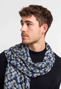 Ung mand med kort krøllet hår iført en marineblå sweater og et blåt tørklæde med hvide blomsterfisker, der ser til venstre.