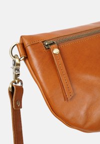 Braune Lederhandtasche mit abgerundeter Form, ausgestattet mit einem Reißverschluss oben, verstellbarem Träger und Messingbeschlägen. Strukturierte Oberfläche mit Steppnähten.