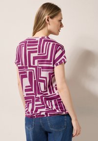 Kortærmet t-shirt i let stof med et geometrisk lilla mønster på en lys baggrund, med en afslappet pasform og synlige sømme.