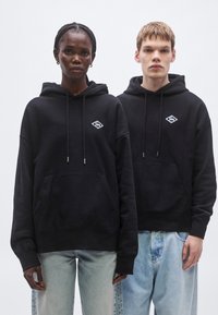 Čierne mikiny s kapucňou majú predné vrecko na brušku, šnúrky a malý diamantový logo na hrudi. Nosené s svetlomodrými džínsami.