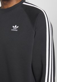 Svart Adidas sweatshirt med vit tre-randig detalj på ärmen och liten vit Adidas trefoil-logotyp på bröstet.