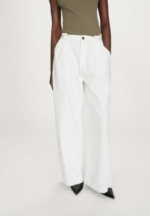 Femme portant un pantalon blanc à taille haute et jambes larges, un haut sans manches vert olive, avec des chaussures noires à bouts pointus, debout les mains dans les poches.