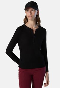 Camisa negra de manga larga con detalle en los botones y escote redondo, combinada con pantalones burdeos y una gorra negra.