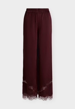 Donna che indossa una canottiera bordeaux con bordo in pizzo e pantaloni coordinati, tiene una clutch nera e si trova davanti a uno sfondo neutro.