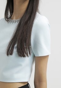 Light blue cropped top with short sleeves, featuring a beaded neckline. Smooth fabric texture and tailored fit.