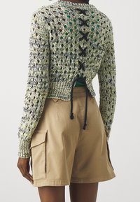 Strickjacke in Grün, Gelb und Marineblau. Kurz geschnittenes Design, offene Rückseite mit Schnürdetails. Kombiniert mit beigen Shorts mit Seitentaschen.