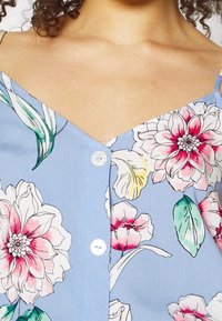 Bloemendruk knoop-overhemd met een lichtblauwe achtergrond. Voorzien van grote roze en witte bloemen, groene bladeren en een V-halsontwerp.