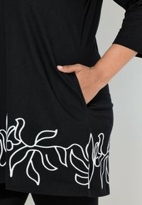 Schwarzes Tunika-Top mit weißer Blumenstickerei entlang des Saums und der Seitentaschen, gefertigt aus weichem Stoff mit lockerem Schnitt.