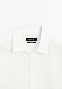 Weißes Hemd mit Kragen und Knopfverschluss, mit glatter Textur und einem Etikett, das "Massimo Dutti" auf schwarzem Hintergrund zeigt.