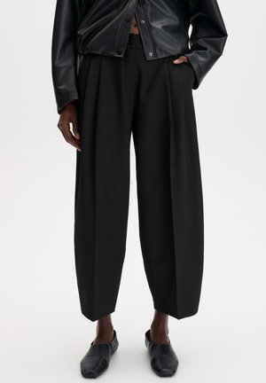 Person wearing loose black cropped trousers, black leather button-up jacket, and black slip-on shoes against a plain background.