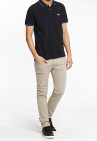 Man i svart pikétröja, beigefärgade slim-fit byxor och svarta sneakers, står med ena handen i fickan mot en vit bakgrund.