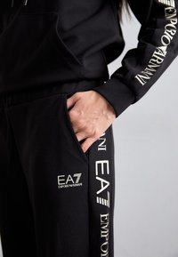Sweat-shirt noir avec impression de logo sur la manche, associé à un pantalon assorti avec une poche avant et des accents de marque en blanc.