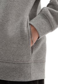 Ruka v kapse šedého svetru s viditelným stříbrným náramkem na zápěstí na bílém pozadí.