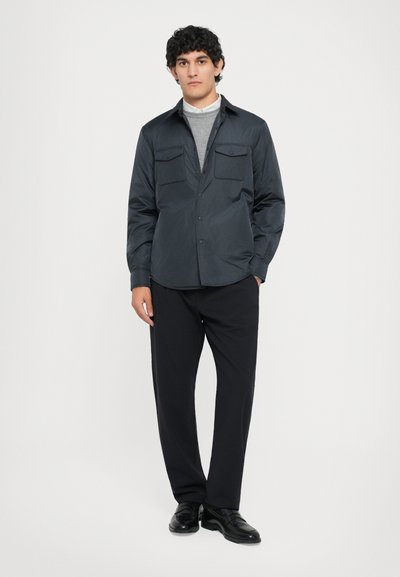Tume sinine särk nööpidega esiküljel, kahe rinna taskuga ja pikkade varrukatega, peal hallist turtleneckist, koos mustade pükste ja kingadega.