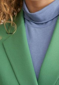 Zielona marynarka z ostrymi klapami na jasnoniebieskim golfie. Tkaniny wydają się gładkie, z wyraźną strukturą i kontrastowymi kolorami.