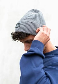 Gray knitted beanie with a ribbed texture and a small black tag. Model adjusts the hat while wearing a dark blue sweatshirt.