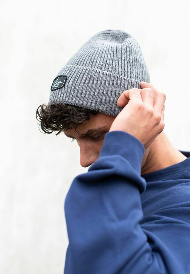Gray knitted beanie with a ribbed texture and a small black tag. Model adjusts the hat while wearing a dark blue sweatshirt.