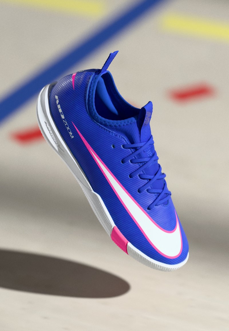 Modré tenisky Nike Air Zoom s bílým a růžovým swoosh, tkaničkami a bílou podrážkou plovoucí nad podlahou tělocvičny s barevnými čarami na hřišti.