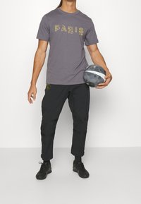 Muž ve šedém tričku s nápisem "PARIS", černých kalhotách a černých teniskách držící černobílý fotbalový míč na bílém pozadí.