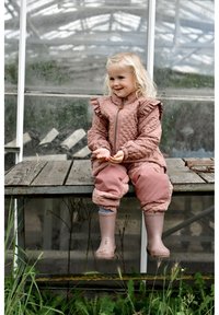 Roosa tikitud laste välisriietus volangidega õlgadel, lühemate käistega, tõmblukuga ja elastsete varrukaservadega. Kandmises heledate kummikute juurde. Taustal puidust pink.