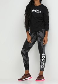 Sweatshirt noir avec le logo "adidas" blanc, leggings camouflage gris avec le texte "adidas" blanc, et baskets marron avec des accents roses.