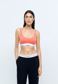 Korall színű sportmellény széles pántokkal, sima textúrával és rugalmas szalaggárral, amelyen Calvin Klein márkajelzés található. Fekete nadrággal párosítva.