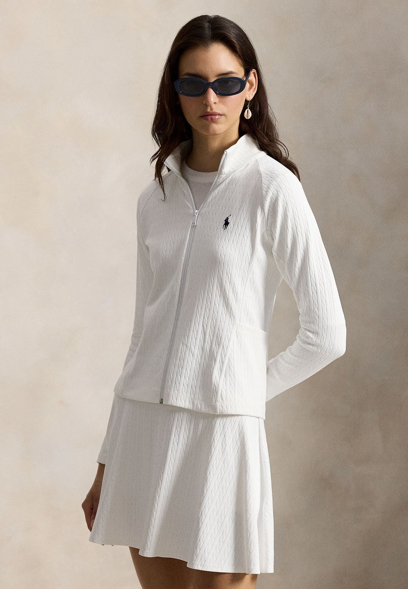 Femme portant une veste blanche texturée à fermeture éclair et une jupe assortie, des lunettes de soleil foncées et une boucle d'oreille en coquillage, debout devant un fond beige.