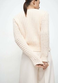Pull tricoté rose clair avec un motif texturé, poignets côtelés et ourlet court, porté sur un pantalon clair, les mains croisées dans le dos.
