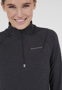 Lächelnde Frau, die ein dunkelgraues Sportoberteil mit Viertelreißverschluss und "ENDURANCE"-Logo auf der Brust trägt, vor einem einfarbigen hellen Hintergrund.