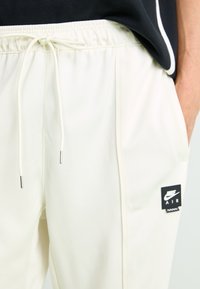 Nærbillede af cremefarvede Nike Air sweatpants med snøre i taljen og sort logo-patch på højre lår, hånd i lommen.