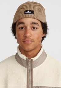 Beige ribbed knit beanie with a black logo patch; warm white fleece jacket with gray zipper and contrasting texture.