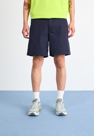 Mørkeblå shorts laget av lett materiale, med rett snitt, elastisk midje og minimale sømdetaljer. Grå sportssko med hvite sokker.