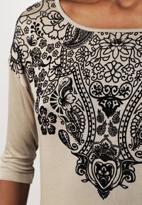 Beige top with three-quarter sleeves featuring intricate black floral and paisley patterns. Soft, textured fabric with a scoop neckline.