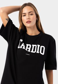 Svart bomull t-shirt med fet vit text "CARDIO", avslappnad passform, korta ärmar och logodetalj nära nederkanten.