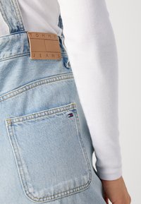 Šviesiai mėlynos džinsinės kombinezonai su rudąja odine etikete, ant kurios parašyta "TOMMY JEANS." Savybės: didelis užpakalinis kišenė ir smulkūs siuvimo detalės.
