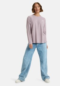 Lange mouwen lavendel T-shirt met een ronde halslijn en een klein logodetail op de borst, gecombineerd met lichtblauwe wijde jeans en witte sneakers.