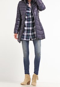 Puffer coat in dark purple, knee-length with quilted design, layered over a plaid shirt in blue and green. Worn with skinny jeans and tan heeled boots.