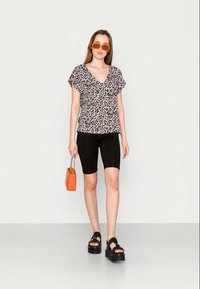 Blusa preta com padrão floral e mangas curtas, combinada com calções de bicicleta pretos. Sandálias de plataforma pretas e uma mala laranja completam o visual.