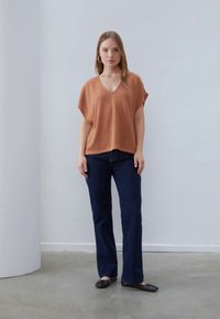 Pletený top v hrdzavej farbe s výstrihom do V a krátkymi širokými rukávmi, kombinovaný s tmavomodrými rozšírenými džínsy a čiernymi plochými topánkami. Jednoduchý dizajn.