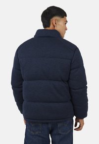 Kurtka puchowa w ciemnym granacie z fakturowanym materiałem, wysokim kołnierzem i poziomym pikowaniem. W połączeniu z niebieskimi dżinsami.